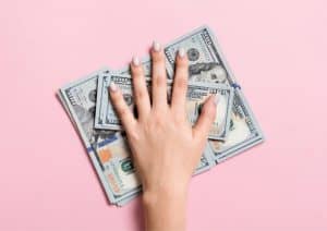 A hand grabbing a stack of 100 dollar bills in front of a pink background