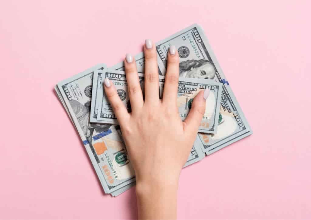 A hand grabbing a stack of 100 dollar bills in front of a pink background